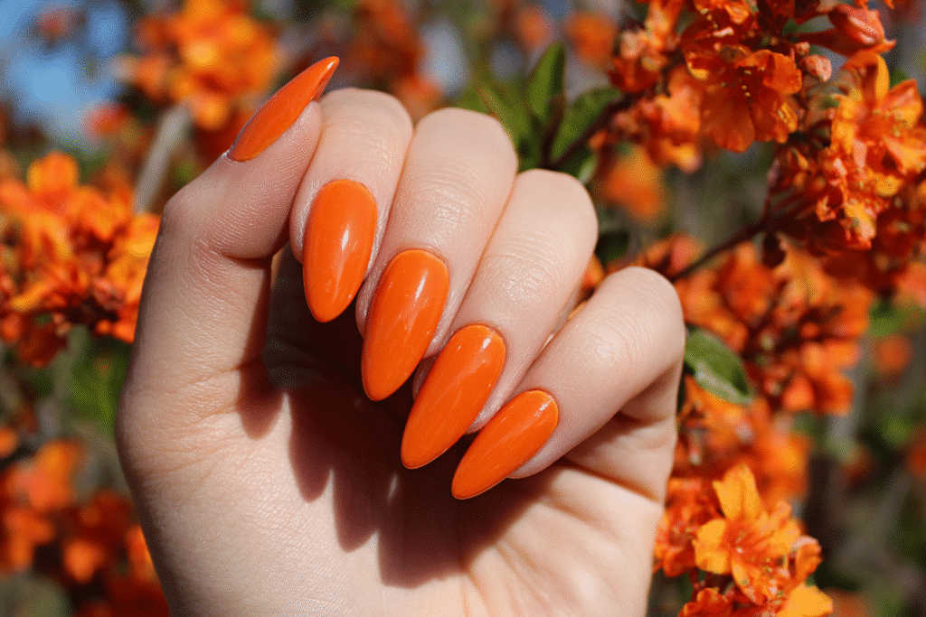 Bold orange summer gel nails with a glossy finish, styled in an almond shape and paired with vibrant orange floral background